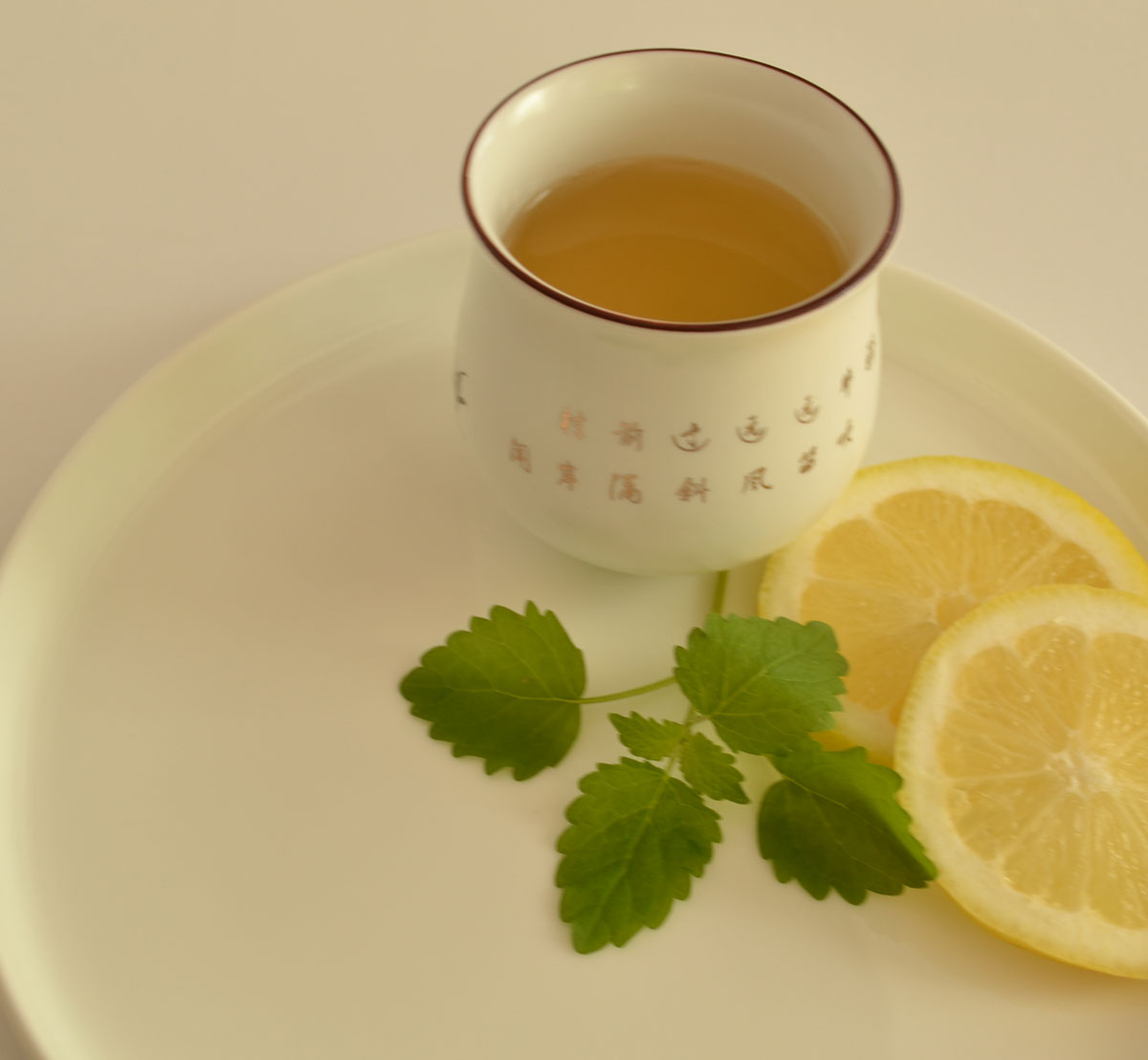 Cup of herbal tea with fresh lemon balm leaves and lemon slices on a white plate.