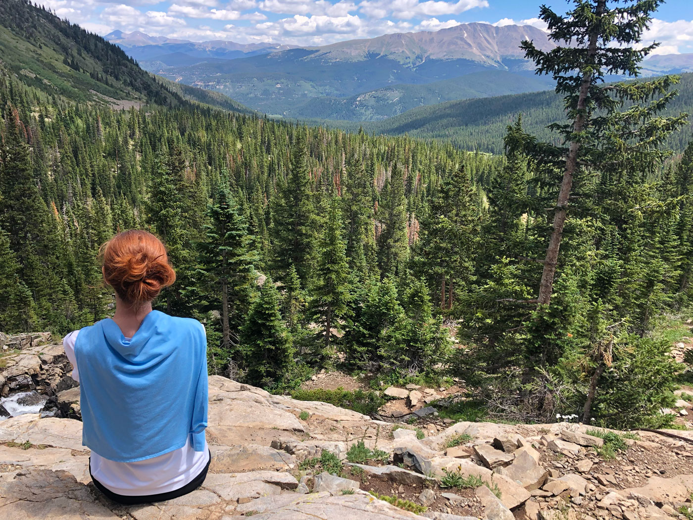 A person with red hair sits on a rocky ledge, looking out over a vast evergreen forest and distant mountains